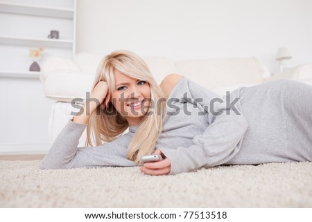 Beautiful blond woman playing with cell phone lying down on a carpet in the living room