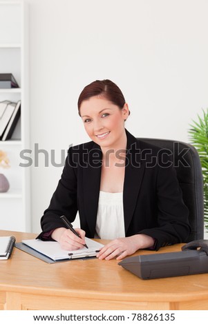 Beautiful red-haired woman in suit writing on a notepad and posing while sitting in an office