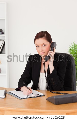 Young gorgeous red-haired woman in suit writing on a notepad and phoning while sitting in an office