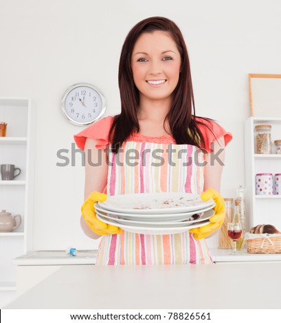 Good looking red-haired woman posing while holding some dirty plates in the kitchen in her apartment