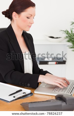 Young attractive red-haired woman in suit typing on her laptop while sitting in an office