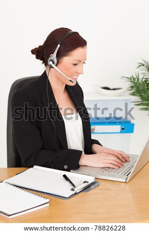 Young attractive red-haired woman in suit typing on her laptop and using headphones while sitting in an office