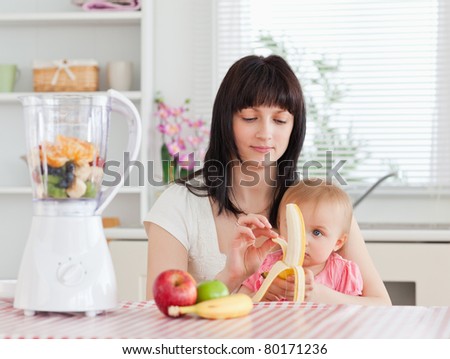 Cute brunette woman pealing a banana while holding her baby on her knees in the kitchen