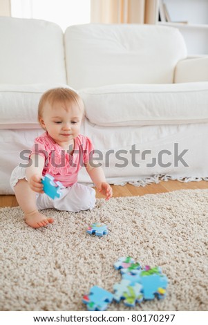 Lovely blond baby playing with puzzle pieces while sitting on a carpet in the living room