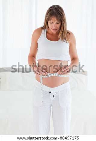 Good looking pregnant female using a tape measure while standing in her bedroom