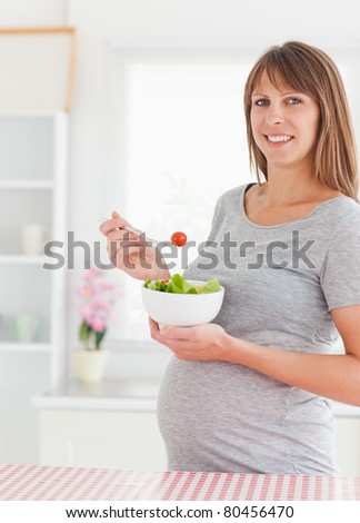Charming pregnant woman eating a cherry tomato while standing in a kitchen