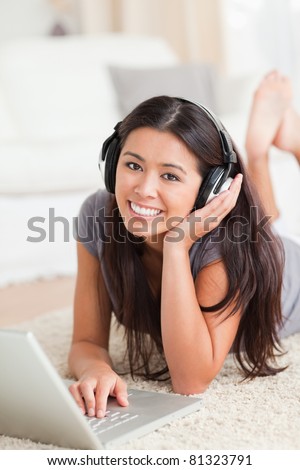 charming woman lying on a carpet with laptop and earphones looking into camera in living room