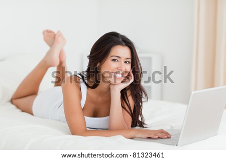 charming woman lying on bed with crossed legs and laptop looking into camera in bedroom