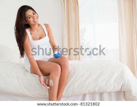 beautiful woman putting creme on her legs while sitting on bed looking into camera in bedroom
