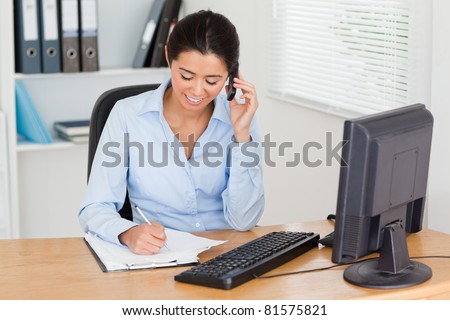 Gorgeous woman using her mobile phone while writing on a sheet of paper at the office