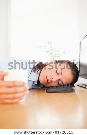 A businesswoman is sleeping in the office with her head in the keyboard while holding a cup of coffee
