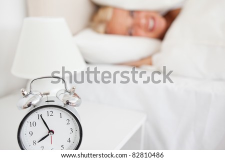 Woman hiding her head in a pillow while the alarm clock is ringing in her bedroom