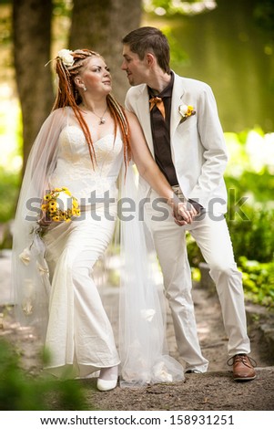 Bride with red dreadlocks and white dress looking at handsome groom in park