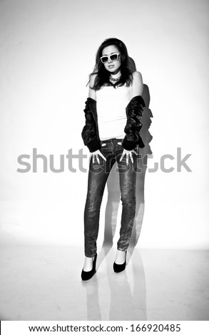 Black and white full length portrait of woman in jeans and leather jacket leaning against white background