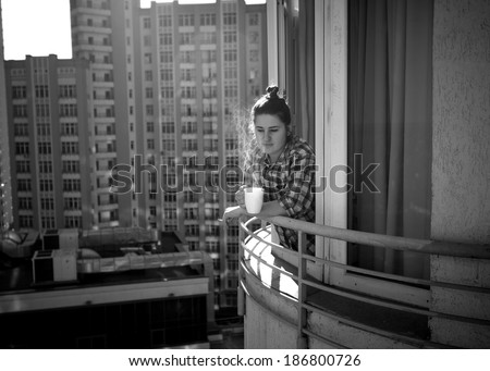 Black and white photo of depressed woman drinking coffee and smoking cigarette on balcony