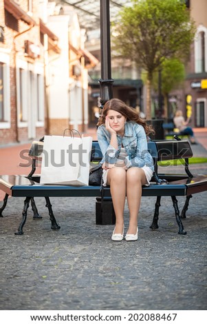 Outdoor photo of sad woman sitting on bench with shopping paper bag