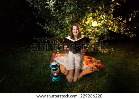 Young smiling woman sitting on grass at night and reading big old book