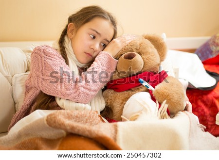 Portrait of little girl holding hand on teddy bears head and measuring temperature