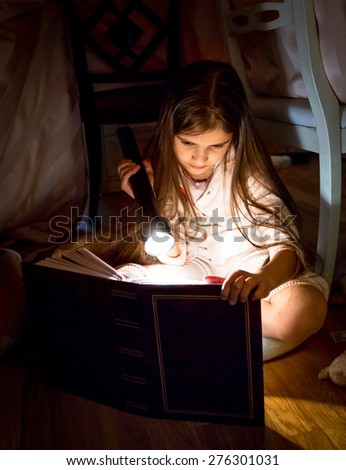 Cute little girl reading book under blanket at night