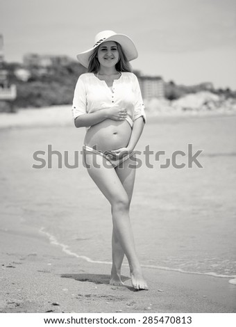 Black and white toned photo of cute pregnant woman in hat posing on sea beach