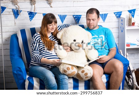 Smiling pregnant woman and husband reading big book to teddy bear on sofa