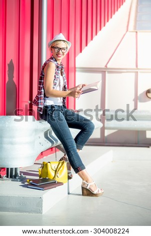 Happy hipster girl sitting on street and holding notebook at hot sunny day