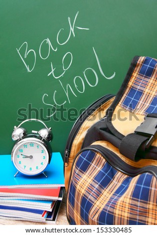 School bag and an alarm clock with writing-books. Against a school board. Back to school.