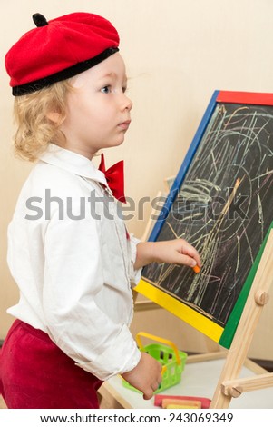 Cute child girl drawing chalk on easel in suit of artist in preschool in kindergarten