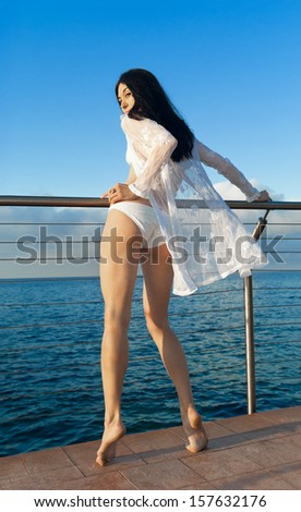 Sexy young brunette woman in white bikini and transparent blouse stands near sea. Sicily. Italy