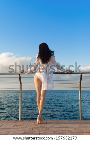 Sexy young brunette woman in white bikini and transparent blouse stands near sea. Sicily. Italy