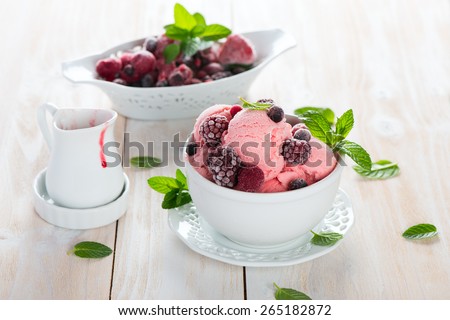 Scoops of pink ice cream in a white bowl, frozen berries, jam and mint on a light wooden surface. Selective focus.