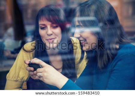 Two young girls smiling using smart phone in a cafe. The photo was taken through the window
