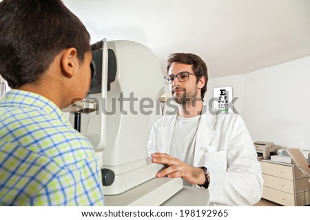 Ophthalmologist In Exam Room With Little Boy Sitting In Chair Looking Into Eye Test Machine