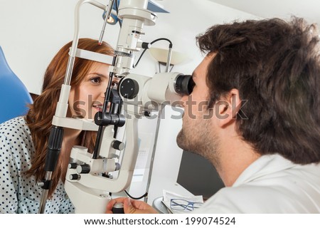 Ophthalmologist In Exam Room With Young Woman Sitting In Chair Looking Into Eye Test Machine