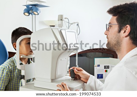 Ophthalmologist In Exam Room With Little Boy Sitting In Chair Looking Into Eye Test Machine