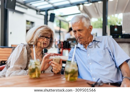 Mature Couple In Cafe Watching Pictures Of Their Grandchildren On Smart Phone