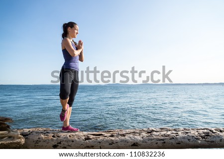 Young women on beach in upright yoga pose with water in background. She is posing in the Tree pose.
