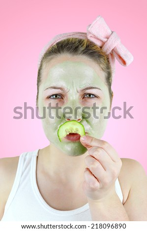 Portrait of a funny woman wearing a pink headband, white top and a green facial mask is holding a cucumber mustache above her lips.