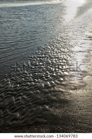 A background of sun glistened, sand ripples and waves on a beach on the ocean.