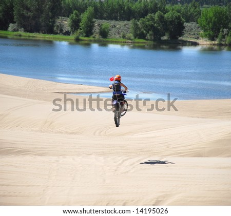 A motorcycle jumps over a sand dune with a lake in the background.
