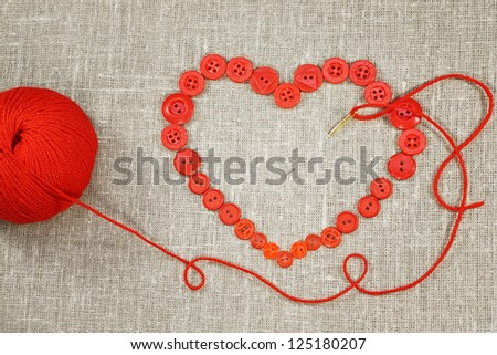 Heart in shape of red buttons, needle and yarn on unbleached fabric.