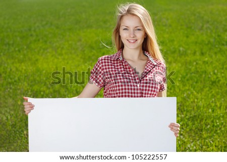 Closeup of young beautiful smiling blond woman holding blank whiteboard, on green background of summer nature