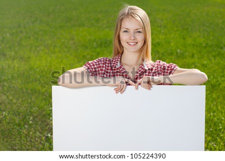 Closeup of young beautiful smiling blond woman holding blank whiteboard, on green background of summer nature