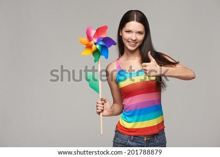 Happy young woman holding toy multicolored pinwheel windmill and gesturing thumb up, over gray background