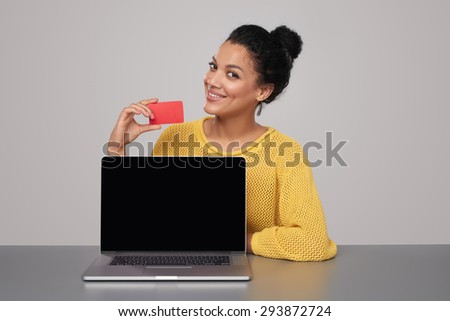 Smiling mixed race african american - caucasian woman showing blank black laptop computer screen and blank credit card, sitting at table over gray background