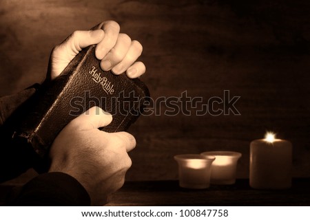 Praying man hands holding and clinching an old holy bible sacred book in a dark church during a prayer worship service with religious candles glowing in nostalgic aged sepia
