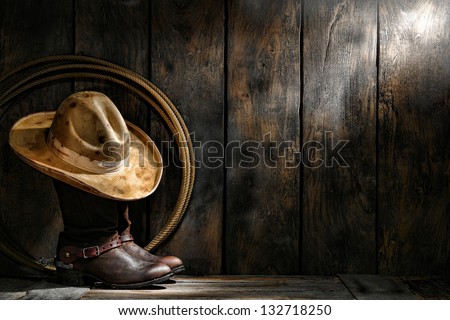 American West rodeo cowboy dirty used felt hat atop worn leather working rancher roper boots with old spurs and lasso lariat in an antique ranch weathered wood cabin