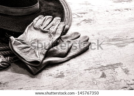American West rodeo worn leather ranching gloves on authentic western cowboy hat atop a vintage lasso lariat over grunge wood planks ranch barn table