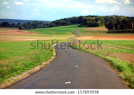 Small narrow country road paved and marked winding with S curves and gentle turns up a hill through the French countryside along farmland and fields in rural France