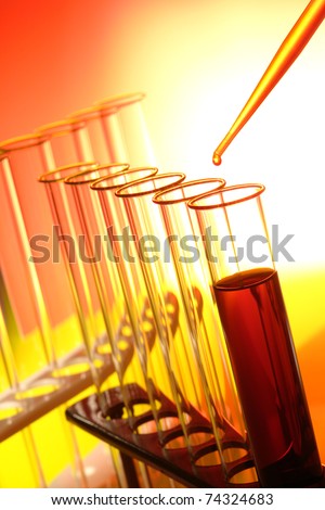 Pipette full of orange liquid with drop of chemical dripping above laboratory test tubes on a rack for a chemistry experiment in a science research lab